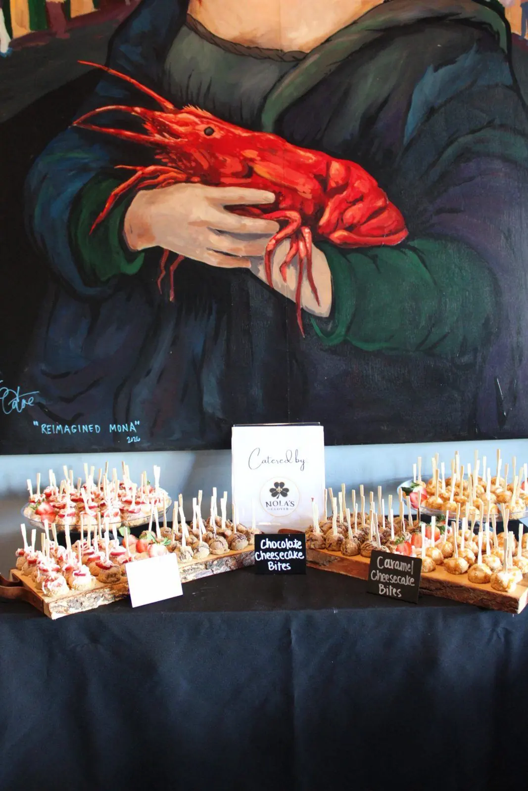 Dessert table of chocolate, caramel, and strawberry cheesecake bites under the Reimagined Mona mural, with a Catered by Nola's Clover sign